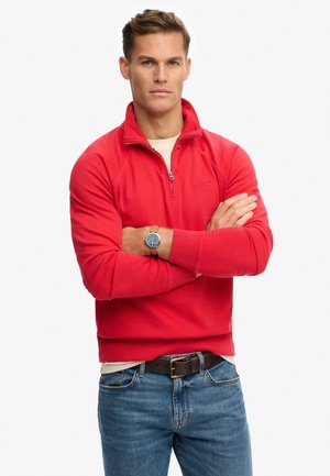 Sweat-shirt rouge à col zippé, avec des poignets côtelés et une texture lisse, associé à un jean bleu et une ceinture marron.