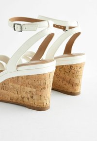 Sandales compensées blanches avec une semelle en liège, dotées de plusieurs brides et d'une boucle à la cheville pour un maintien sûr. Texture lisse et design épuré.