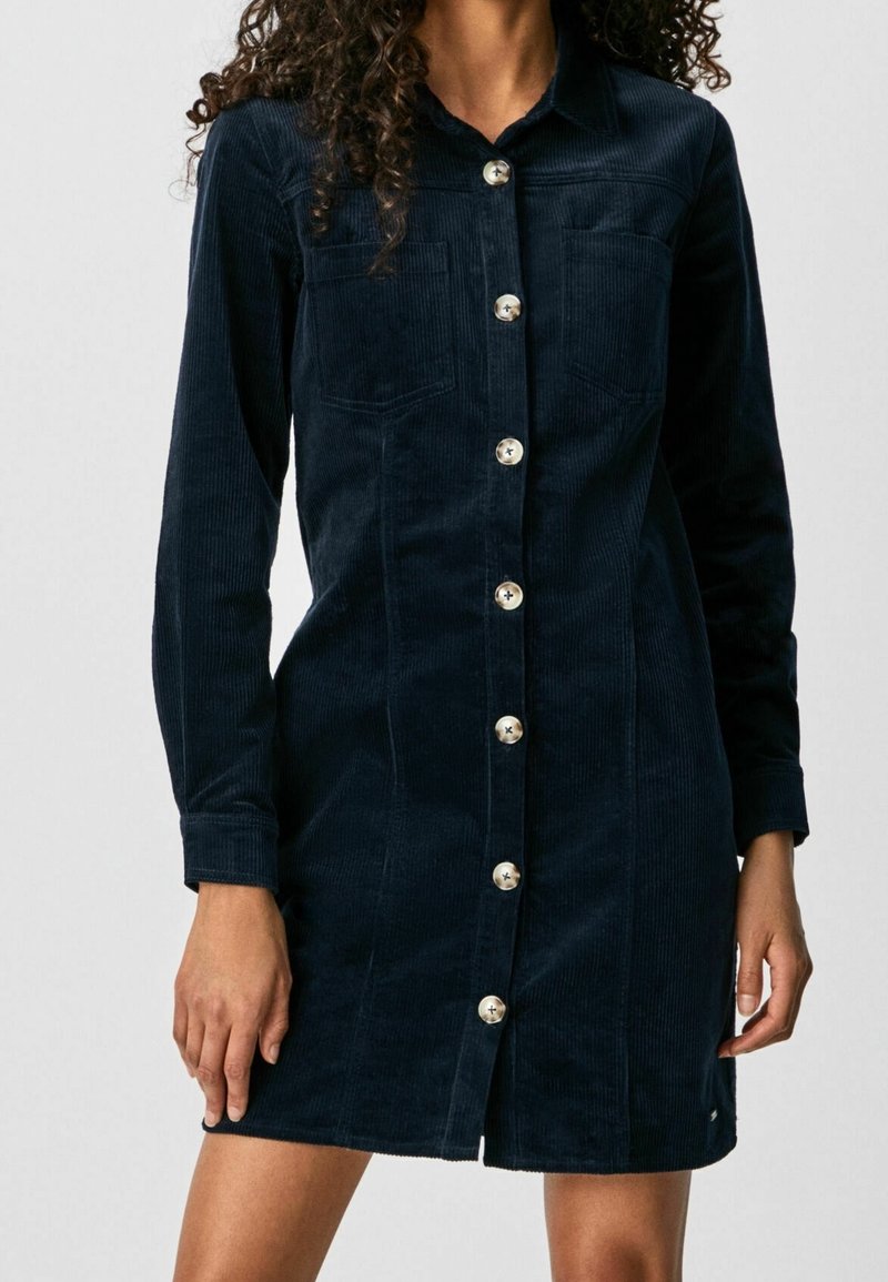 Tummansininen samettinen nappimekko, jossa on pitkät hihat, etutaskut ja kaulus, jota kantaa seisova henkilö.