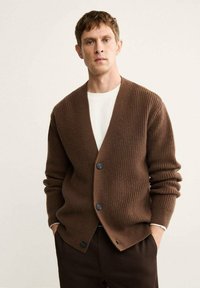 Cardigan marrone a coste con scollo a V profondo e tre bottoni, realizzato in un materiale a maglia testurizzata. Indossato sopra una camicia bianca.