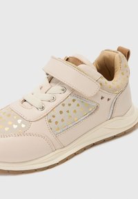 Baskets beiges avec une tige en cuir texturé, des accents argentés et une sangle Velcro. Présente une section à motifs avec de petits cœurs en or.