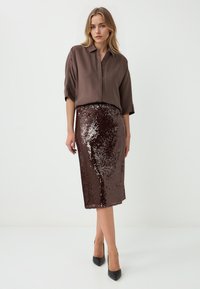 Braune Bluse mit lockerer Passform und kurzen Ärmeln, kombiniert mit einem knielangen, mit Pailletten besetzten Rock in Dunkelbraun. Schwarze Absatzschuhe runden das Outfit ab.