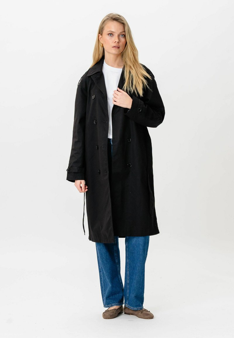 Femme blonde portant un long manteau noir à double boutonnage, une chemise blanche, un jean bleu et des chaussures plates marron, debout devant un fond blanc.