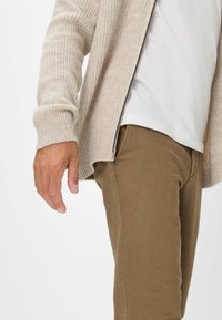 Beige geribde cardigan met een rits en een wit T-shirt eronder, gecombineerd met bruine broeken. Opmerkelijke details zijn een knoop op de broeken.