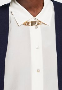 Marinblå stickad cardigan över en krämfärgad blus, med en knapplistdesign, ett kedjehalsband med guldtonade detaljer och en mjuk textur.