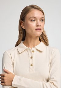 Polo shirt a maniche lunghe di colore crema con una placca di bottoni caratterizzata da bottoni dorati, colletto piatto e dettaglio del logo ricamato sulla manica.
