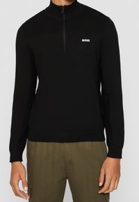 Black knit quarter-zip pullover with a high collar and textured upper panel. Features a white "BOSS" logo on the chest.