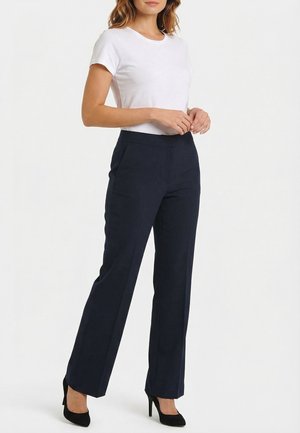 Mujer con top blanco de manga corta, pantalones azul marino de pierna recta y zapatos de tacón negro, de pie con las manos entrelazadas.