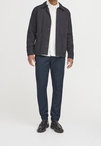 Svart bomullsjacka med framfickor, knappstängning och krage, lagd över en vit skjorta. Mörkblå slim-fit byxor och svarta skor.