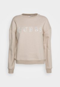 Beige sweatshirt lavet af blødt stof, med rund hals, drop-skuldrer og "GUESS" logo i skinnende bogstaver på forsiden.