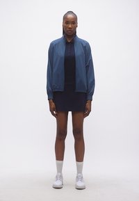 Γυναίκα που στέκεται όρθια φορώντας μπλε σκούρο σακάκι, σκούρο φόρεμα πόλο, λευκές κάλτσες και λευκά αθλητικά παπούτσια μπροστά σε ένα απλό λευκό φόντο.