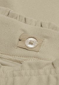 Beige kangas, joka on sileäpintainen ja jossa on röyhelöreuna sekä läpinäkyvä napin yksityiskohta ommellussa nauhassa.