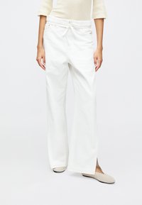 Hvit vide bukser laget av myk stoff, med strikk i midjen, sidelommer og en avslappet passform. Et par myke beige leiligheter synlige.