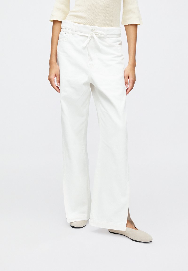 Hvit vide bukser laget av myk stoff, med strikk i midjen, sidelommer og en avslappet passform. Et par myke beige leiligheter synlige.