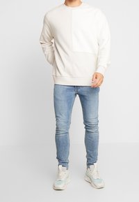 Hombre vestido con un sudadera beige clara, jeans ajustados de color azul claro y zapatillas blancas con acentos en azul claro, de pie contra un fondo blanco.