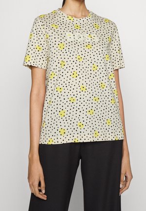 Vrouw draagt een beige T-shirt met korte mouwen met zwarte polka dots, gele vlekken en de tekst "BOSS", gecombineerd met een zwarte broek.