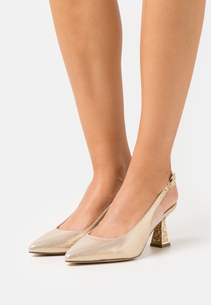 Pieds portant des talons slingback dorés à bout pointu avec un talon bloc présentant des détails texturés et scintillants.