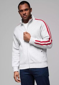Felpa grigia con zip e strisce rosse e bianche sulle spalle, caratterizzata da un colletto alto e polsini a coste, abbinata a jeans scuri.