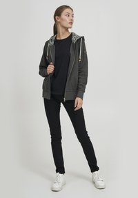 Felpa grigia con zip e fodera a motivo, cappuccio regolabile con cordini e dettagli in legno. Indossata con una maglietta nera e jeans neri, abbinata a sneaker bianche.
