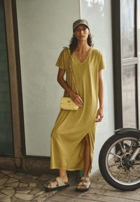 Olijfgroen maxi-jurk met een V-hals, korte mouwen en een split aan de zijkant; gecombineerd met beige schoeisel en een lichtgele schoudertas.