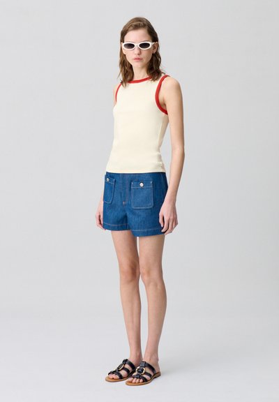 Canotta crema con bordi rossi abbinata a pantaloni corti di jeans blu con tasche frontali. Sandali neri con dettagli in oro.