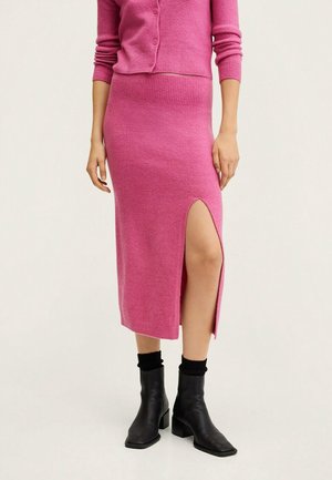 Kobieta ubrana w różową dzianinową spódnicę midi z rozcięciem sięgającym uda, dopasowany kardigan, czarne skarpetki oraz czarne botki na obcasie.