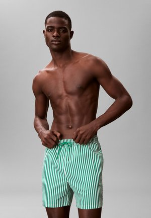 Mężczyzna bez koszulki, dopasowujący zielono-białe spodenki kąpielowe w pionowe paski na jednolitym szarym tle.