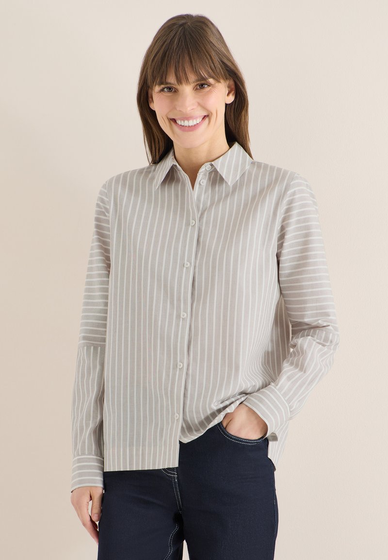 Femme souriante portant une chemise boutonnée à rayures verticales beige et blanche, avec un pantalon bleu foncé, debout avec une main dans la poche devant un fond uni.