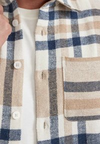 Koszula flanelowa w kratę w kolorach beżowym, niebieskim i białym. Posiada guziki, kieszeń na piersi oraz miękką fakturę z wzorem w kratkę.