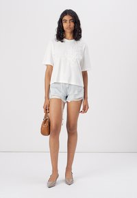 T-shirt court en coton blanc avec broderies florales, associé à un short en denim déchiré. Les accessoires comprennent un petit sac à main marron et des talons gris.