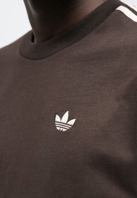 Καφέ βαμβακερό t-shirt με στρογγυλή λαιμόκοψη και διακριτικό σχέδιο με τρεις ρίγες. Ένα λευκό κεντητό λογότυπο στο στήθος.