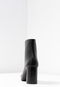 Bottine en cuir noir avec un talon bloc haut, texture lisse, bout rond et détails de couture à l'arrière. Design minimaliste.