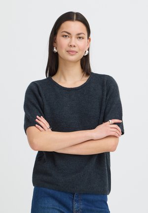 Kvinde med langt mørkt hår iført en mørk kortærmet sweater, sølv øreringe, ringe og blå jeans, stående med armene over kors.