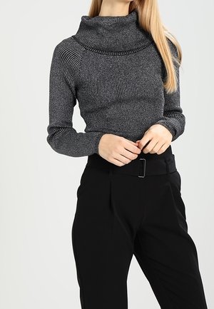 Femme portant un pull col montant en maille côtelée gris foncé et un pantalon noir taille haute avec une ceinture, les mains jointes à la taille.
