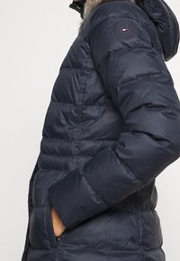 Chaqueta acolchada azul marino con diseño acolchado horizontal y capucha forrada de piel. Cuenta con un parche con el logotipo en el hombro y bolsillos laterales con cremallera.