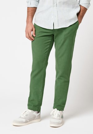 Homme portant un pantalon vert, des baskets blanches et une chemise légère à boutons, les mains dans les poches, sur un fond uni.