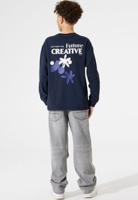 Marineblauwe longsleeve t-shirt met witte en paarse grafische tekst en abstracte vormen op de achterkant, gecombineerd met lichtgrijze jeans en witte sneakers.