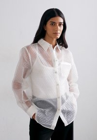 Camicia traslucida con texture a bolle, colletto, tasca frontale e chiusura con bottoni, indossata sopra un top grigio a strati, abbinata a pantaloni neri.