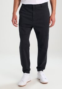 Black tapered pants with elastic cuffs, crafted from cotton, featuring contrasting stitching and a button closure. Worn with white sneakers.