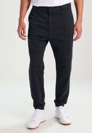 Black tapered pants with elastic cuffs, crafted from cotton, featuring contrasting stitching and a button closure. Worn with white sneakers.