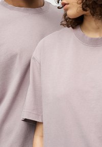 Δύο άτομα φορούν απαλά, ανοιχτό μωβ, υπερμεγέθη βαμβακερά μπλουζάκια με κοντά μανίκια και ριμπ λαιμόκοψη.