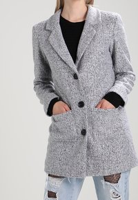 Šedý textured kabát s klopovým límcem, dvěma předními kapsami a třemi knoflíky. Nosený přes černý top a seprané světle modré džíny.