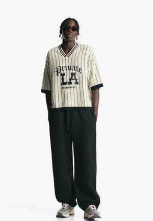 Maglietta oversize a righe crema con dettagli in blu navy, con stampa "Private LA Tournament". Indossata con pantaloni neri larghi e sneaker grigie.