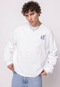 White long-sleeve T-shirt made of cotton, featuring a small graphic on the left chest. Model wears light blue denim, with a chain necklace.