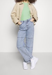 Giacca beige corta sopra un top verde a costine, abbinata a jeans larghi blu chiaro con cuciture orizzontali. Sneakers bianche completano l'outfit.