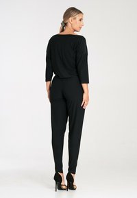 Czarny kombinezon o luźnym fasonie, z rękawami o długości trzy czwarte, zwężanymi nogawkami oraz niskim dekoltem z tyłu, w połączeniu z czarnymi szpilkami.