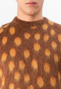 Close-up van een persoon die een fuzzy bruine trui draagt met grote fuzzy geel-oranje stippen en een ronde halslijn.