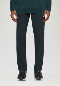 Donkergroene geribbelde trui gedragen met strak gesneden donkere broek en zwarte sportschoenen, die een gladde textuur en strakke lijnen laat zien.