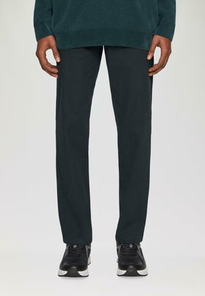 Dunkelgrüner gerippter Pullover, getragen mit gerade geschnittenen dunklen Hosen und schwarzen Sportschuhen, die eine glatte Textur und klare Linien zeigen.