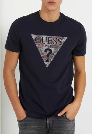 Uomo che indossa una maglietta blu navy con un grande logo triangolare Guess e un punto interrogativo stampato sul davanti, mani nelle tasche.